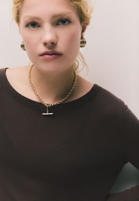 Auksinė grandinėlė su cilindro formos pakabuku, derinama su dideliais auksiniais lankstiniais auskarais. Modelis dėvi tamsų ribuotą viršų su apvalia iškirpte.