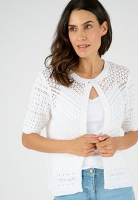 Witte gebreide cardigan met korte mouwen, een textuur with opengewerkt ontwerp en een centrale knoopsluiting. Gepaarde met een wit topje en blauwe jeans.