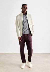 Pull zippé côtelé beige clair, sweat-shirt col rond gris, pantalon bordeaux et baskets blanches ; accessoirisé avec des lunettes de soleil dorées.