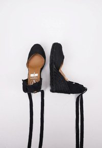Zapatos estilo espadrille en negro, con un tacón de cuña tejido y texturizado, cintas en el tobillo y punta redondeada, que presentan una plantilla beige con marca.