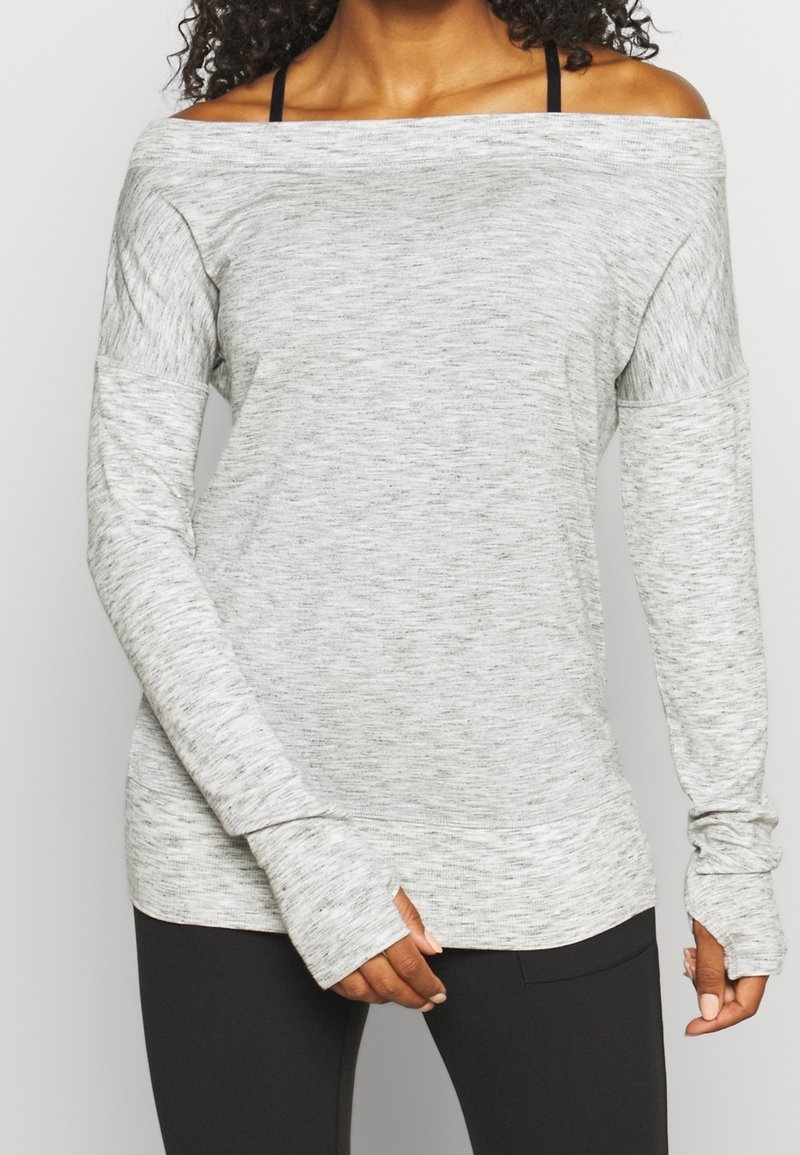 Femme portant un haut gris clair à manches longues et épaules dénudées, ainsi que des leggings noirs, avec des trous pour le pouce dans les manches et des bretelles de soutien-gorge noires visibles.