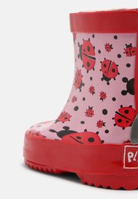 Roter Gummiregenstiefel vor pinkem Hintergrund, mit einem Marienkäfer und schwarzem Punktmuster. Mit strukturierter Sohle und Markenetikett.