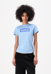Šviesiai mėlynas trumpomis rankov�ėmis marškinėliai su mėlynu stačiakampiu "HUGO" logotipu per krūtinę; lygus paviršius; derinami su juodomis kelnėmis.
