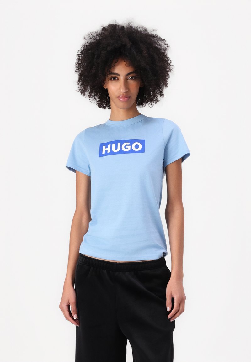 Šviesiai mėlynas trumpomis rankovėmis marškinėliai su mėlynu stačiakampiu "HUGO" logotipu per krūtinę; lygus paviršius; derinami su juodomis kelnėmis.