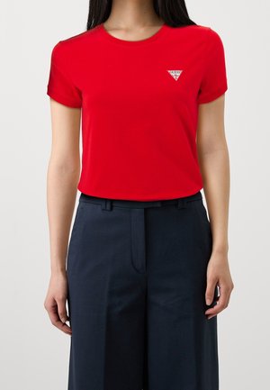 Vrouw draagt een rode T-shirt met korte mouwen en een klein wit Guess-logo op de borst, gecombineerd met een donkere marineblauwe broek met hoge taille.
