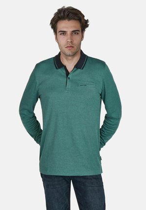 Grünes langärmeliges Polo-Shirt mit strukturierter Webart, schwarzem Kragen, Drei-Knopf-Leiste und kleinem Brustlogo; kombiniert mit blauen Jeans.