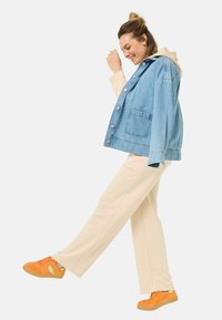Leren spijkerjack in lichtblauw met zilveren knopen, gedragen over een beige hoodie. Gecombineerd met crèmekleurige broeken en levendig oranje sneakers.