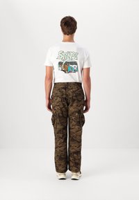 Hvid grafisk t-shirt med grønt "STANCE" logo og illustreret varevogn på ryggen, parret med brune camouflage cargo bukser med sidelommer.
