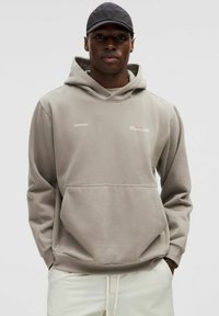 Sweat à capuche en tissu gris clair avec un design simple, doté d'une poche avant et d'un détail de logo sur la poitrine.