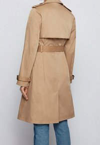 Beżowy płaszcz trencz z paskiem w talii, rozcięciem z tyłu i guzikami na pagonach na ramionach. Wykonany z gładkiej, wodoodpornej tkaniny.