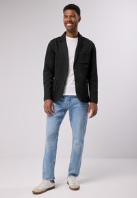 Černý blazer z pevné tkaniny s klopovými límci, s jednou kapsou; pod tím bílý tričko. Světle modré džíny a bílé tenisky doplňují outfit.