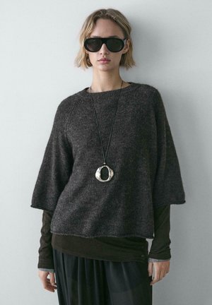 Maglione marrone scuro, dalla vestibilità ampia con maniche larghe e una finitura texture. Completato da una collana argento circolare spessa e occhiali da sole neri.
