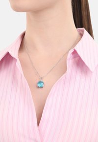Collana d'argento con un pendente quadrato, di colore blu chiaro e sfaccettato. Indossata su una camicia a righe rosa con colletto, che mette in risalto la chiarezza e la forma del pendente.