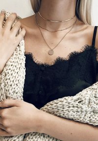 Czarna koronkowa koszulka bez rękawów z frędzlami, nałożona z trzema delikatnymi naszyjnikami. Gruby, kremowy sweter jest zarzucony na ramiona.