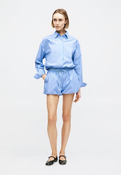 Γυναίκα στέκεται φορώντας ανοιχτό μπλε ριγέ πουκάμισο με μακριά μανίκια, κρυμμένο μέσα σε ταιριαστά σορτς με κορδόνι, μαύρα παπούτσια Mary Jane, με τα χέρια στις τσέπες.