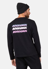 Schwarzes Langarm-T-Shirt mit mehrfarbigem "ROSSIGNOL"-Logo, das in gestapeltem Text auf dem Rücken gedruckt ist. Kombiniert mit schwarzen Hosen und einer Strickmütze.