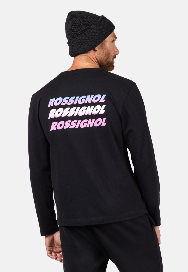 Schwarzes Langarm-T-Shirt mit mehrfarbigem "ROSSIGNOL"-Logo, das in gestapeltem Text auf dem Rücken gedruckt ist. Kombiniert mit schwarzen Hosen und einer Strickmütze.