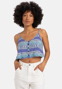 Niebieski i fioletowy wzorzysty crop top z regulowanymi ramiączkami, z guzikami z przodu i geometrycznymi wzorami. Noszony z białymi spodniami o wysokim stanie.