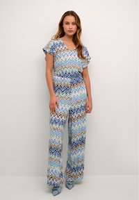 Mitmevärviline ühesuunaline kombekas, millel on sinise, kreemi ja pruuni toonide zigzag muster; lühikesed lendavad varrukad, V-kaelus ja laiad jalad.