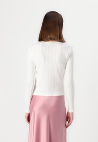 Haut à manches longues blanc avec une texture côtelée et un motif subtil, associé à une jupe rose lisse. La vue de dos montre un décolleté courbé.