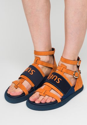 Piedi con sandali aperti arancioni e blu navy con cinturini alla caviglia e fibbie su uno sfondo chiaro.