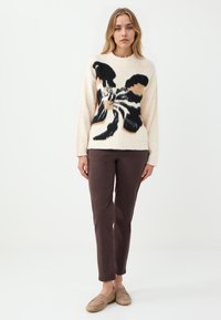 Cremefarbener Pullover mit schwarzem und beigem Schmetterlingsdesign, kombiniert mit braunen Hosen und beigen Slippern, die weiche Texturen aufweisen.