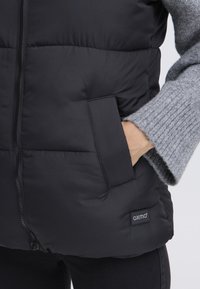 Zwart gewatteerd bodywarmer met doorgestikt ontwerp, ritssluiting aan de voorkant, zijzakken en een "OXMO" logo-patch. Het model draagt daaronder een grijze geribde trui.