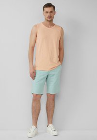Pfirsichfarbene Tanktop und hellblaue Shorts, beide aus leichtem Material. Das Model trägt weiße Sneakers und steht vor einem neutralen Hintergrund.