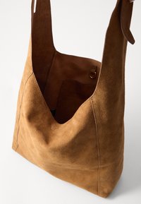 Brown suede shoulder bag with a slouchy design, prominent stitching, an interior pocket, and a wide shoulder strap. Smooth texture.