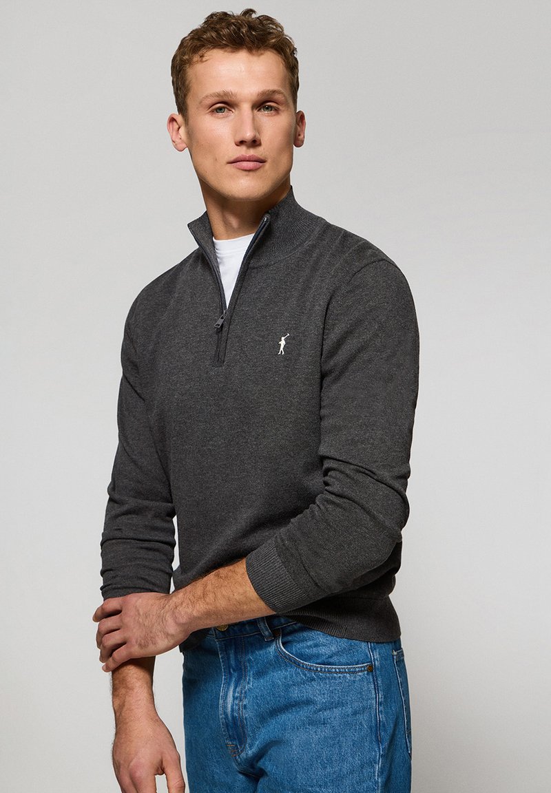 Suéter gris con cremallera fabricado en tejido de punto suave, con un alto cuello y puños acanalados. Pequeño logo ubicado en el lado izquierdo del pecho.
