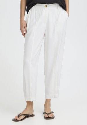 Kobieta ubrana w białe spodnie typu tapered z mankietami, czarną bluzkę i ciemnobrązowe płaskie sandały typu thong, stojąca z rękami w kieszeniach.