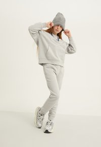 Gaiši pelēks sporta krekls ar kabatu, saskaņots ar atbilstošām sporta biksēm. Aksesuārs ir teksturēta pelēka cepure un sudraba sporta apavi.