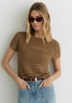 Kvinde iført brun strikket kortærmet top med udskårne mønstre, blå denim bukser, mørke solbriller og et mørkebrunt bælte med guldspænde.