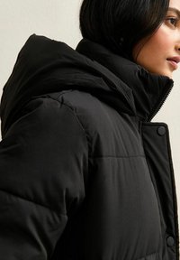 Giacca imbottita nera con colletto alto e grande cappuccio. Presenta una zip frontale e bottoni a pressione, mostrando un design trapuntato e testurizzato.