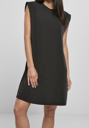 Nainen pukeutuneena hihattomaan, mustaan polvipituiseen mekkoon, jossa on jäsennellyt olkapäätyynyt, seisomassa yksinkertaista harmaata taustaa vasten.