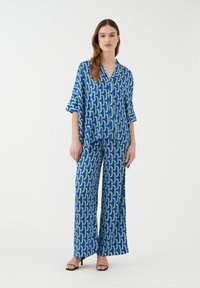 Tenue assortie composée d'un chemisier ample à manches courtes et d'un pantalon large. Tissu bleu avec un motif géométrique vert et noir. Texture lisse.