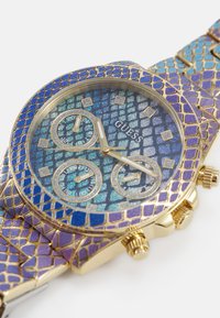 Montre dorée avec un cadran texturé en motif peau de serpent bleu et violet. Dispose de trois sous-cadrans, d'aiguilles dorées et d'un bracelet texturé.