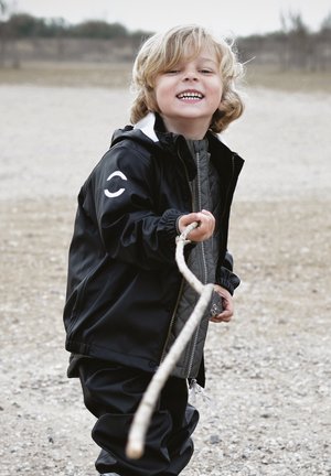 Fekete vízálló dzseki fehér díszítésekkel és steppelt szürke belső réteggel, hozzáillő nadrággal. Egy gyerek egy botot tart a kezében.
