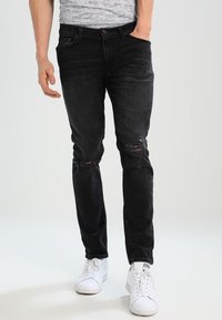 Jeans de mezclilla negra con detalles desgastados, corte ajustado y textura descolorida, combinados con zapatillas blancas. El modelo se encuentra de pie de forma casual con las manos en los bolsillos.