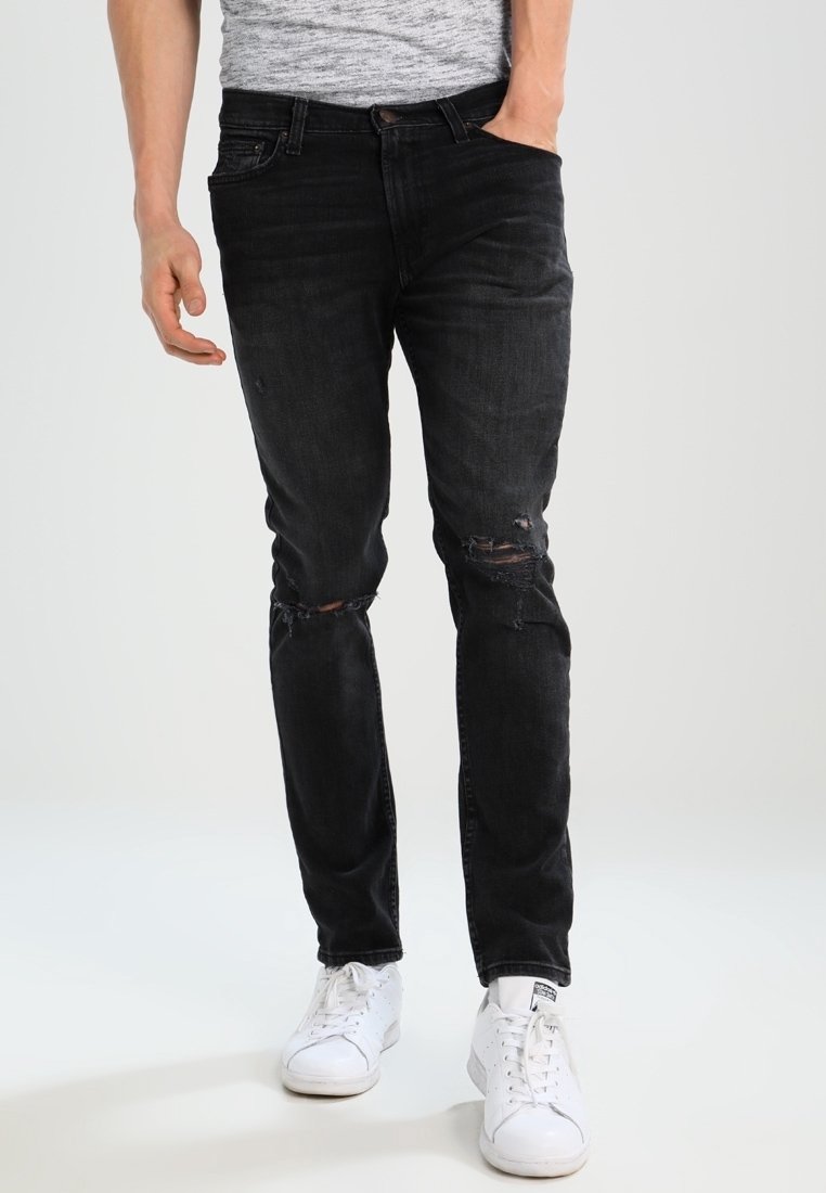 Jeans de mezclilla negra con detalles desgastados, corte ajustado y textura descolorida, combinados con zapatillas blancas. El modelo se encuentra de pie de forma casual con las manos en los bolsillos.