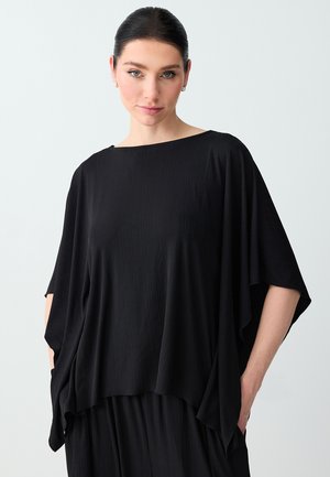 Kobieta o ciemnych włosach w luźnej, czarnej, krótkim rękawie, teksturowanej bluzce i pasujących szerokich spodniach, stojąca na tle jasnego, jednolitego tła.