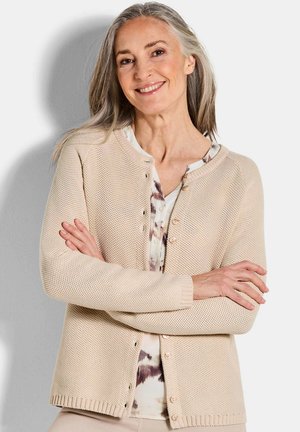 Oudere vrouw met grijs haar glimlacht, draagt een beige gebreide cardigan over een gebloemde blouse, met gekruiste armen, staand tegen een effen achtergrond.
