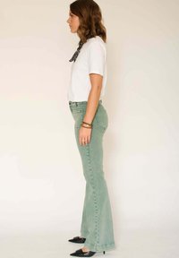 Jean évasé vert clair à taille haute, associé à une chemise blanche à manches courtes et des escarpins noirs à bouts pointus. Écharpe à motifs autour du cou.