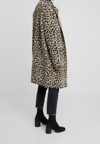 Płaszcz w panterkę o miękkiej teksturze, w odcieniach beżu i czerni, zestawiony z czarnymi zamszowymi botkami za kostkę oraz ciemnoniebieskimi dżinsami.