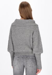 Grå stickad tröja med bred krage, strukturerat mönster och ribbade ärmslut. Kombinerad med grå jeans, vilket ger en avslappnad passform.