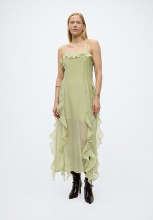 Femme portant une robe sans manches vert clair à volants avec des bretelles spaghetti et des bottines à talons noires, debout devant un fond blanc uni.