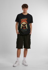 Sort T-shirt med farverig kraniet grafik og tekst, parret med mørke cargo shorts. Model iført hvide sneakers og ankelstrømper.