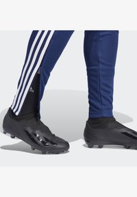 Schwarze Fußballschuhe mit strukturiertem Obermaterial, die drei markante Streifen an der Seite aufweisen. Marineblaue Sporthosen mit Reißverschluss und weißen Akzenten.