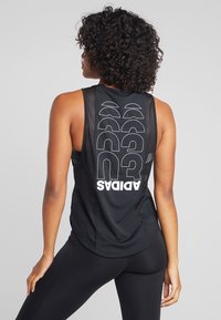 Canottiera sportiva nera con design a rete, schiena allungata e testo bianco "ADIDAS" con grafiche stilizzate disposte in verticale.