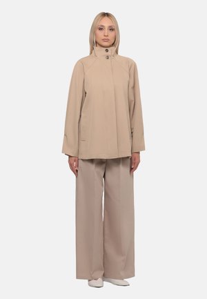 Donna bionda in piedi che indossa una giacca beige con collo alto e bottoni e pantaloni larghi abbinati su uno sfondo chiaro e uniforme.